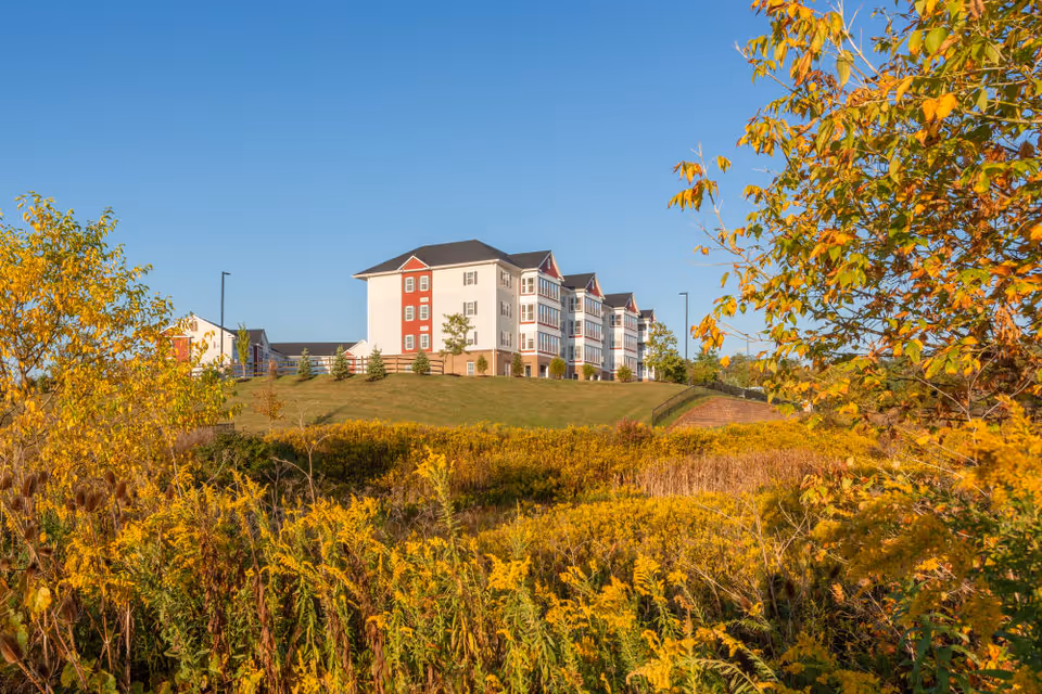 A multi-story senior living facility building situated on a grassy hill surrounded by autumn foliage with yellow and orange leaves under a clear blue sky.