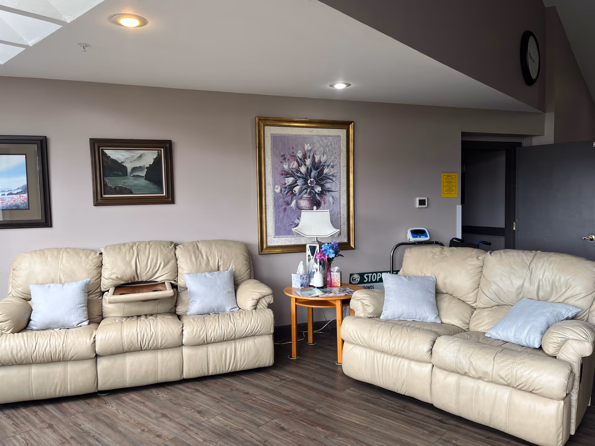 A cozy living area with two beige leather sofas, throw pillows, framed wall art, and a small round side table holding a lamp and flowers.