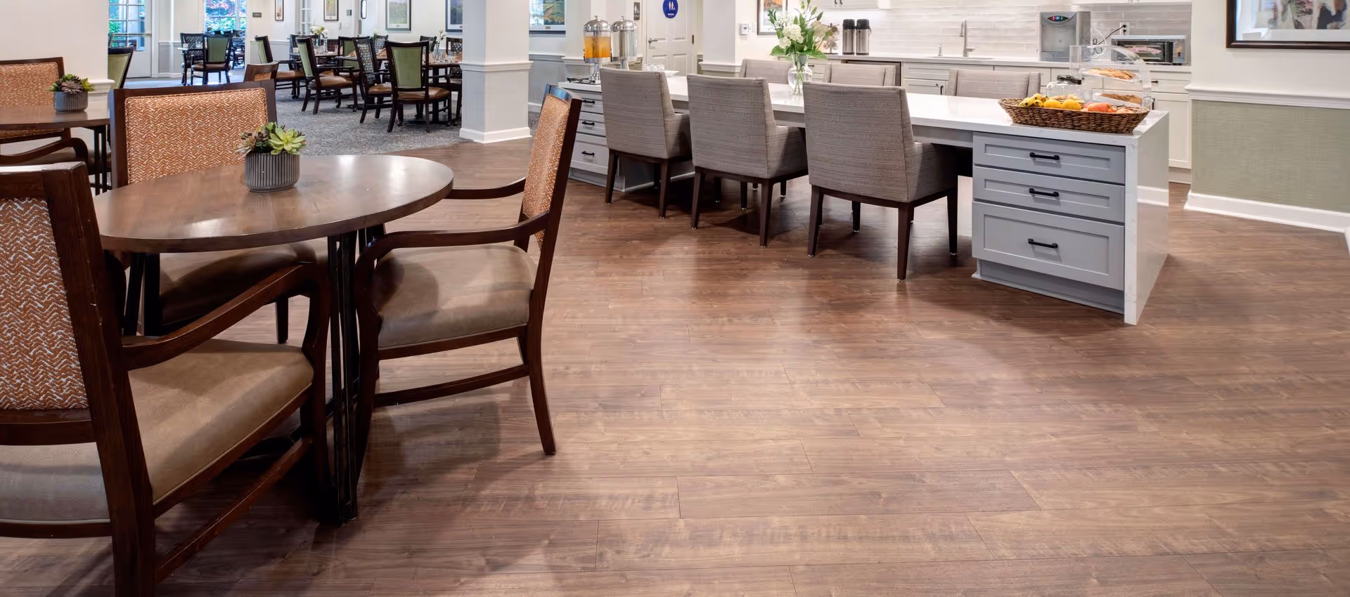 Open dining area with round wooden tables and chairs and a kitchen island with bar stools in a senior living facility.
