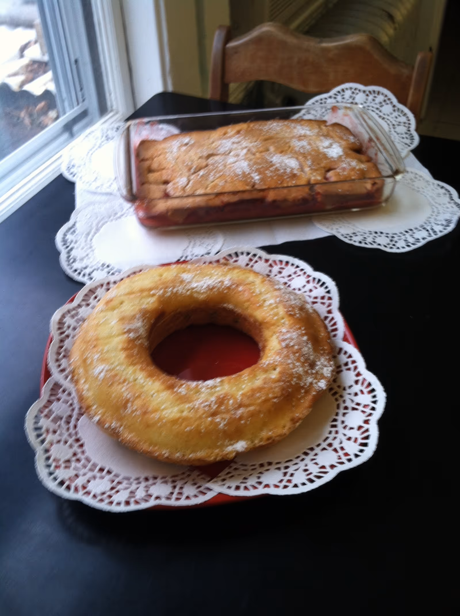 A bundt-style cake and a rectangular fruit cobbler on doilies atop a table by a window.