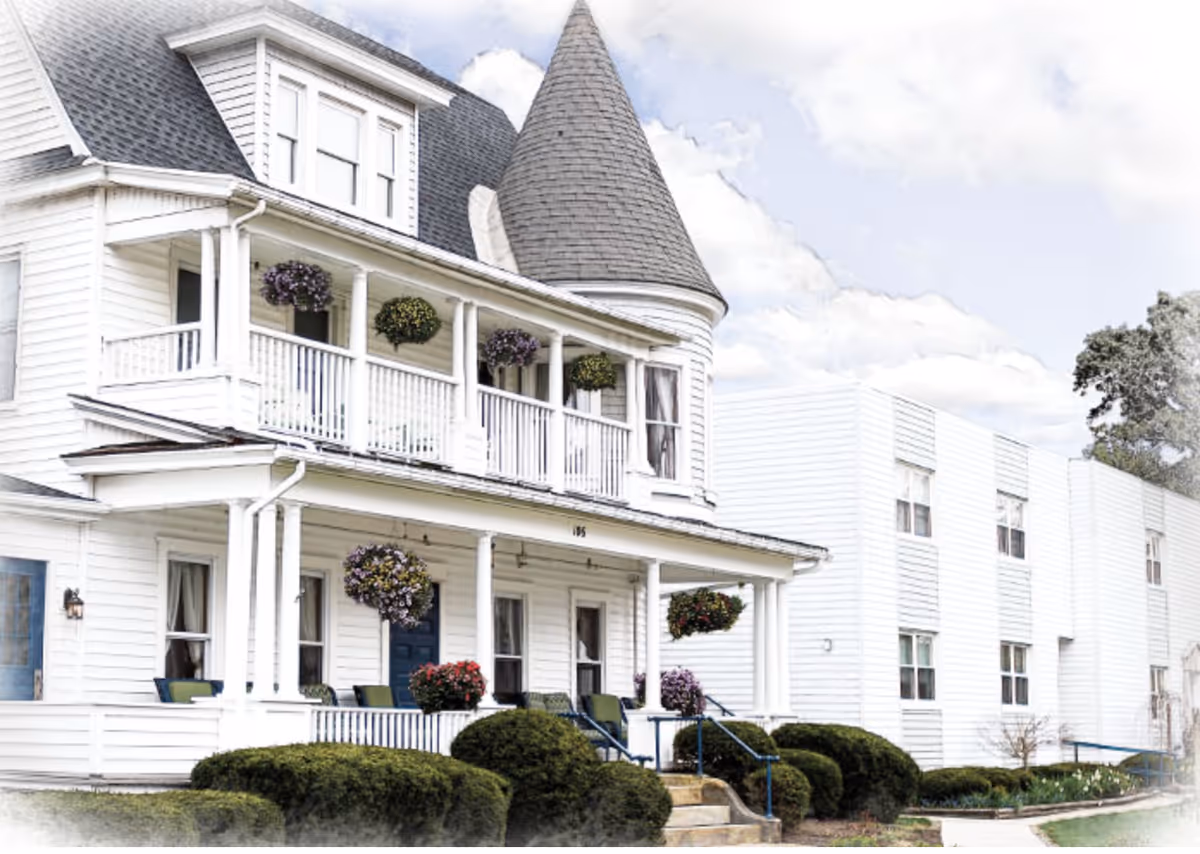 Exterior view of a senior living facility named The Magnolia featuring a white two-story building with a turret, a covered porch with hanging flower baskets, and well-maintained bushes and landscaping.