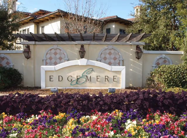 Entrance sign for Edgemere facility with a decorative wall, lantern-style lights, and a colorful flower bed in front. Trees and part of the building are visible in the background.