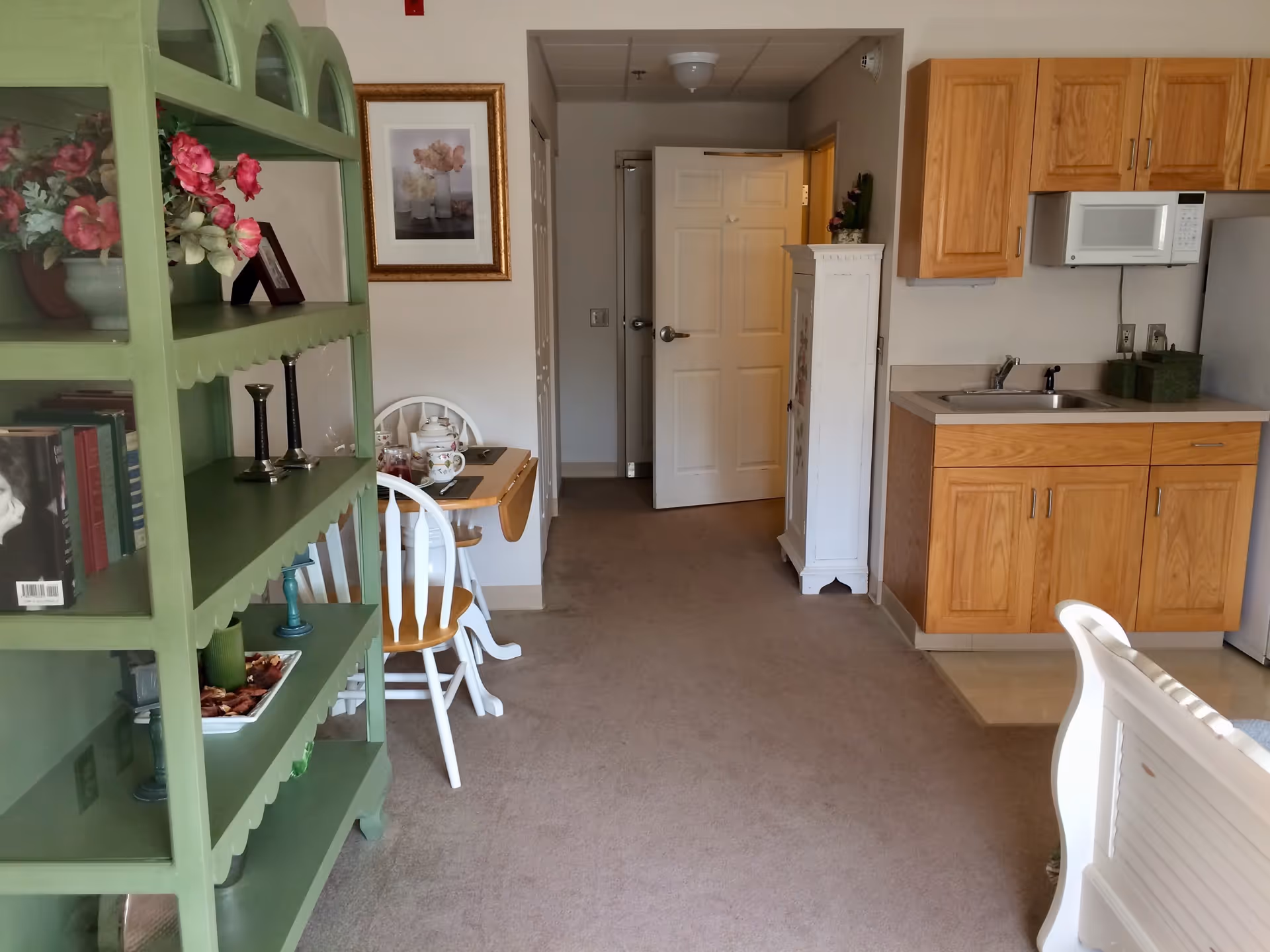 Interior view of a senior living facility room showing a small kitchenette with wooden cabinets, a microwave, and a sink on the right. A green shelving unit with decorative items and books is on the left. A small dining table with two white chairs is placed against the wall near a framed floral picture. The room has beige carpet and a white door at the end of the hallway is partially open.