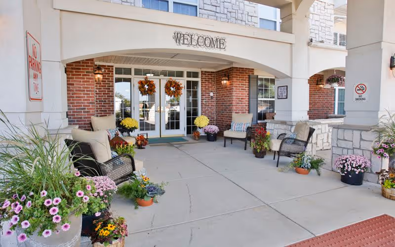 Entrance area of a senior living facility with a covered porch featuring a 'WELCOME' sign above double glass doors. The porch is decorated with fall wreaths on the doors, potted flowers, and cushioned chairs arranged on either side. There are 'No Parking Any Time' and 'No Smoking' signs visible on the pillars.