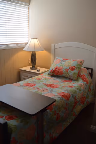 A small bedroom with a single bed covered in a floral bedspread and matching pillow. Next to the bed is a white nightstand with a lamp and a small bell. A window with closed blinds is on the left wall, allowing some natural light into the room.