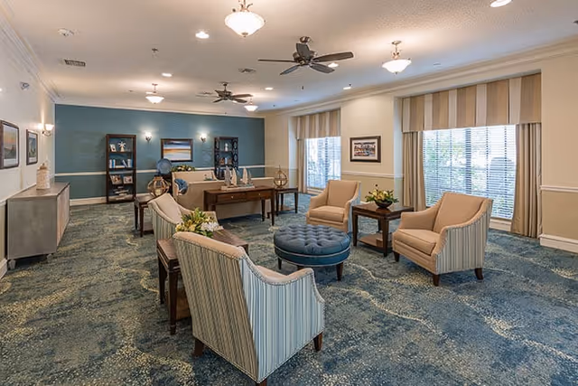 Bright communal living room with several upholstered armchairs, a round tufted ottoman, desks, bookshelves, and large windows.