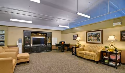 Carpeted common lounge with a sofa, armchairs, TV entertainment center, lamps and a small table.