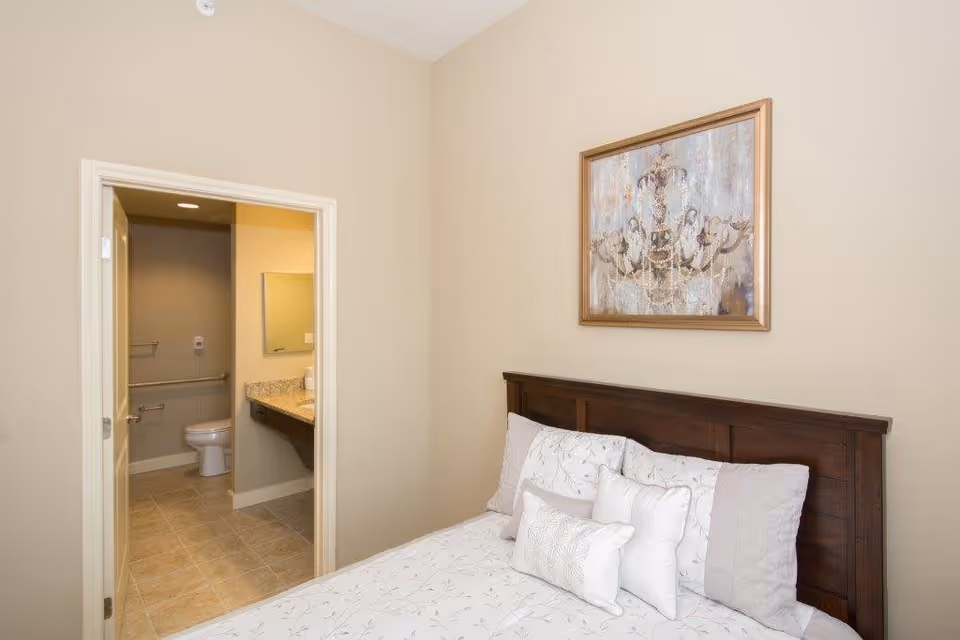 A bedroom with a wooden headboard, decorative pillows and bedding, wall art above the bed, and an open door revealing an adjacent bathroom.