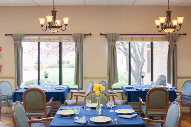 Bright dining room with tables set with blue tablecloths, place settings, chairs, chandeliers, and large windows looking out to a lawn.