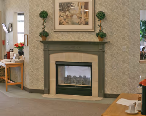 Decorative sitting area with a fireplace mantel topped by a framed painting and two potted topiary plants, with tables visible at the sides.