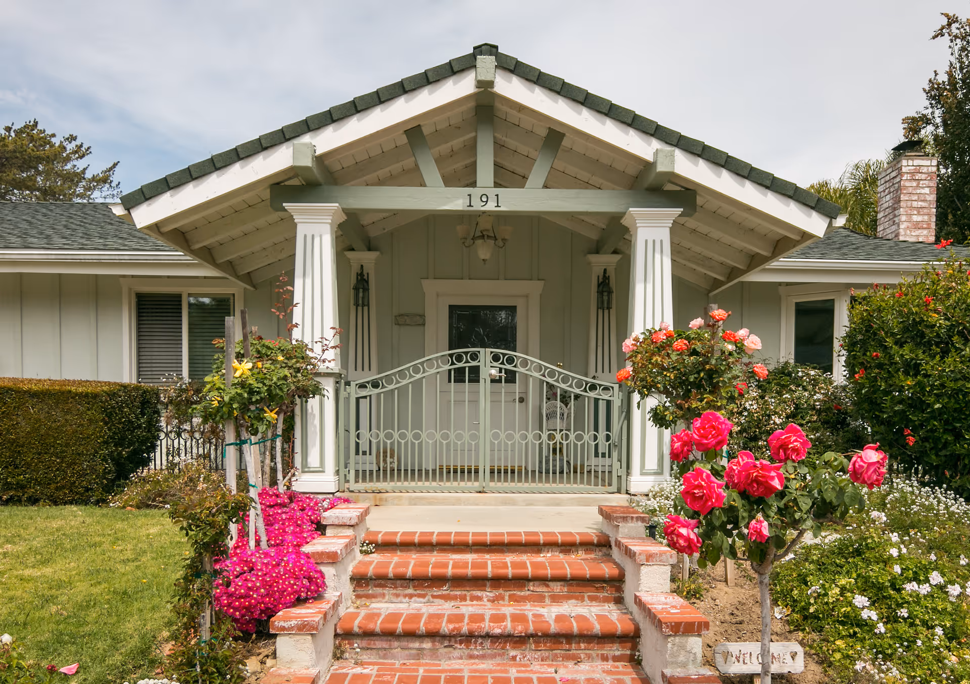 Front entrance of a single-story house with a covered porch supported by white columns. The porch has a decorative metal gate and a white door. Brick steps lead up to the porch, flanked by vibrant pink and red flowers and green shrubs. The house number 191 is displayed above the porch.