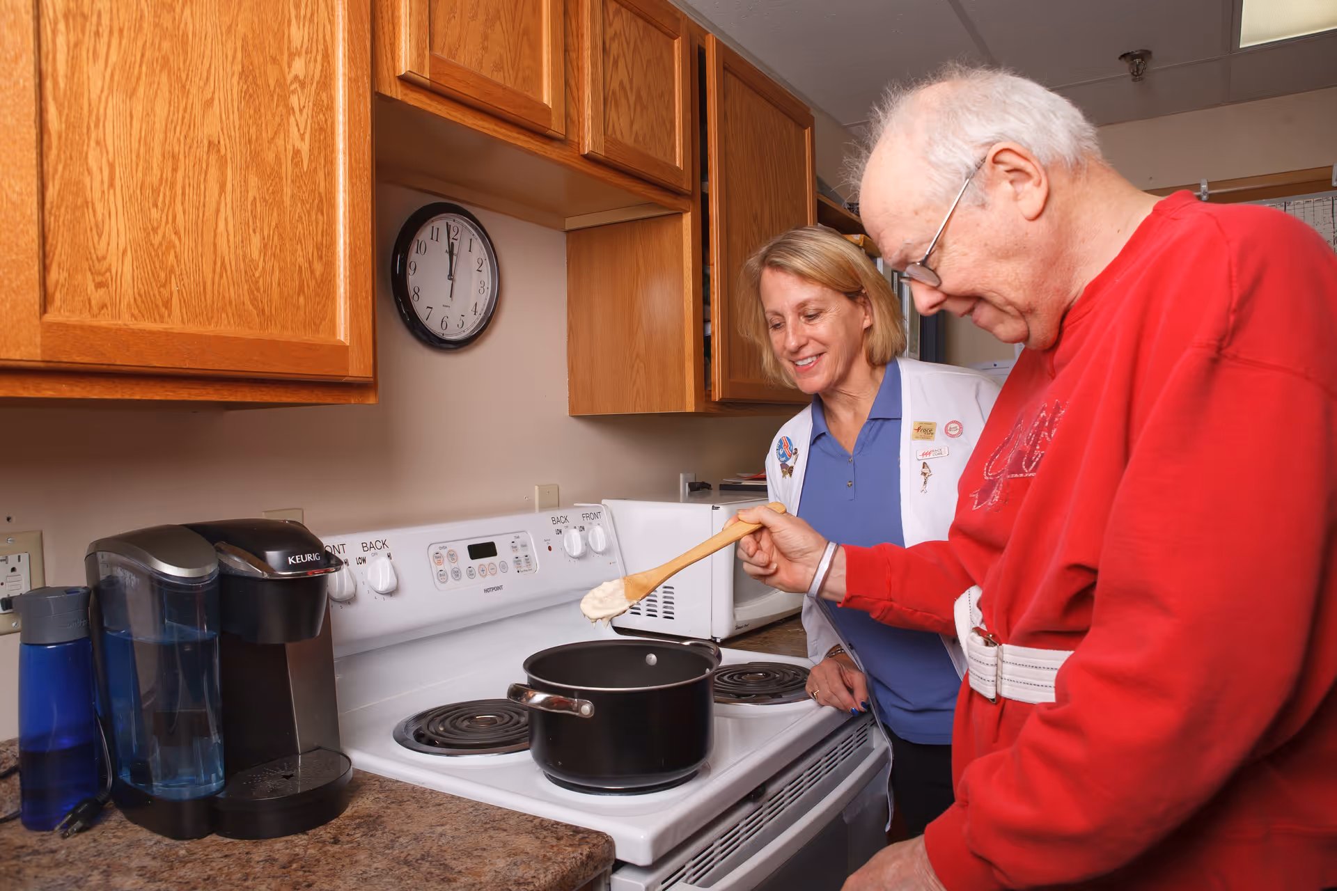 An elderly man stirs a pot on a kitchen stove while a caregiver watches and smiles.