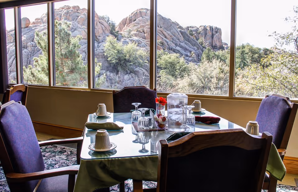 A dining table set with cups, glasses and a water pitcher in a dining room with large windows overlooking rocky cliffs and trees.