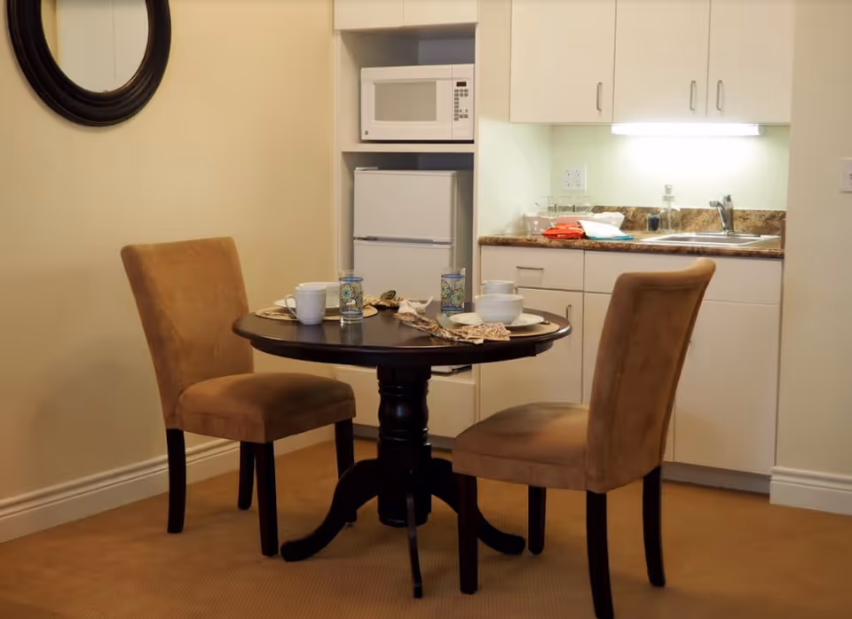 Small kitchenette and dining area with a round table set for two, two upholstered chairs, a microwave, mini fridge, and sink.