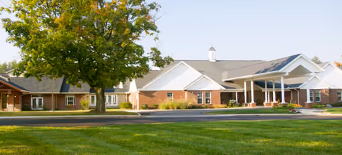 Single-story brick building with a covered entrance, green lawn, and a large tree in front.