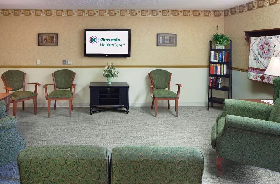 A furnished senior living common room with green upholstered chairs, a wall-mounted TV showing the Genesis HealthCare logo, a small table with flowers, and a bookshelf.