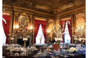 An opulent dining room with ornate gold-trimmed walls and ceiling, red velvet curtains, and large windows. The room is set up for a formal event with round tables covered in dark blue tablecloths, elegant floral centerpieces, and neatly arranged chairs.