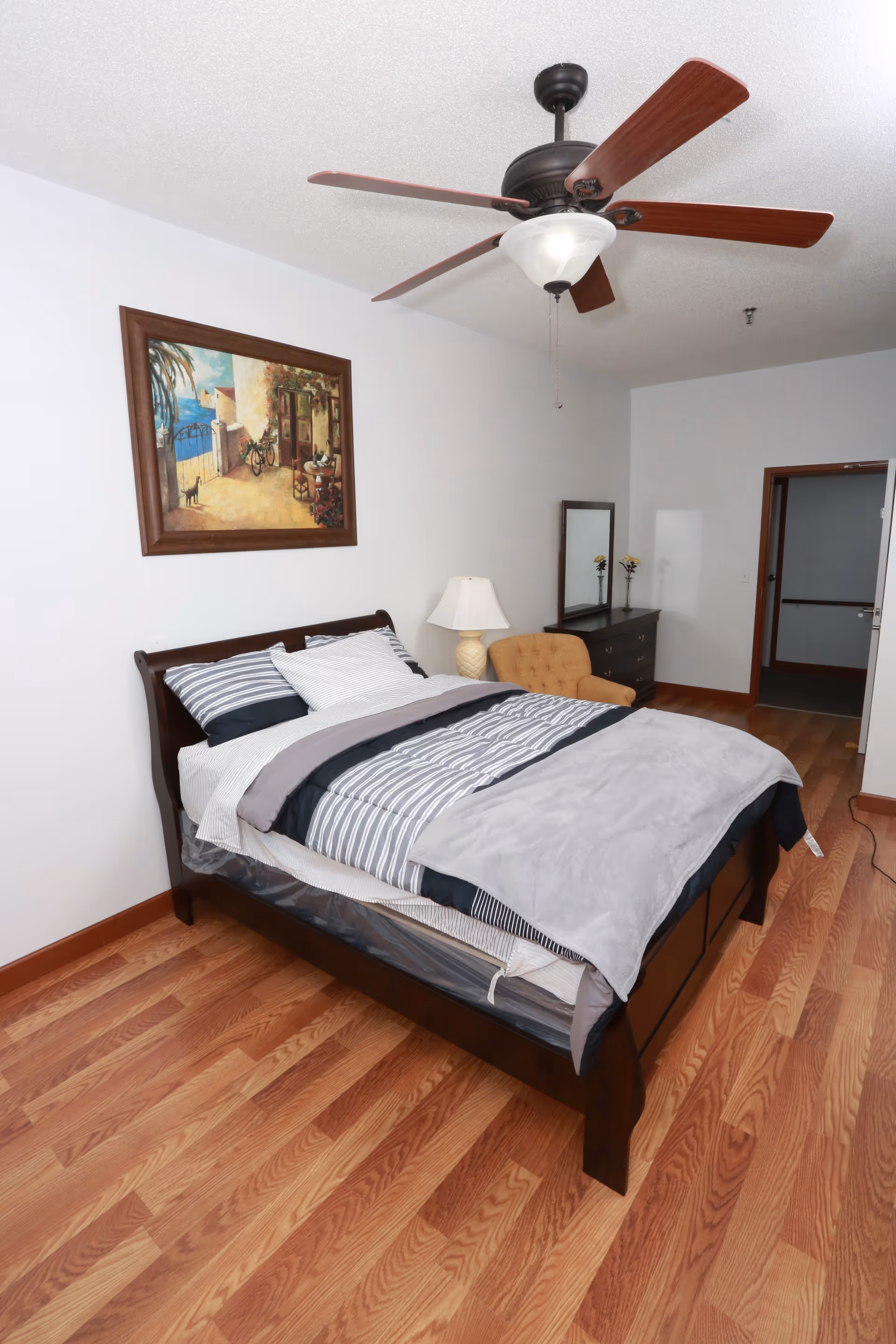 A bedroom with a wooden bed frame, neatly made with striped bedding and a gray blanket. There is a ceiling fan with wooden blades above the bed. On the wall above the bed hangs a framed painting depicting an outdoor scene with a bicycle and a cat. Next to the bed is a nightstand with a lamp, and further back is a dresser with a mirror and two small flower vases. The floor is wooden, and there is an open doorway leading to another room.