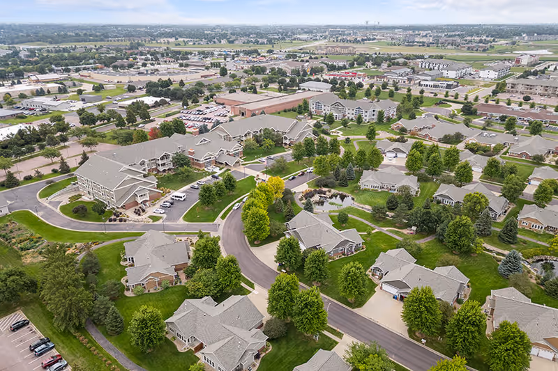 Aerial view of Good Samaritan Society - Prairie Creek facility showing multiple residential buildings surrounded by green lawns and trees, with roads and parking areas connecting the buildings. The surrounding area includes more buildings and open spaces extending into the distance under a cloudy sky.