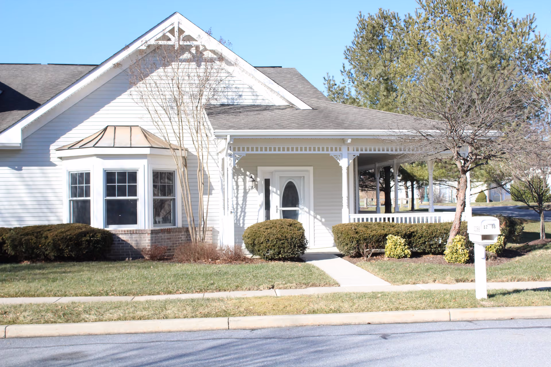 Exterior view of a single-story residential building with white siding, a gray shingled roof, a bay window, and a covered porch. There are trimmed bushes and small trees in the front yard, a sidewalk leading to the front door, and a white mailbox near the street. The sky is clear and blue.