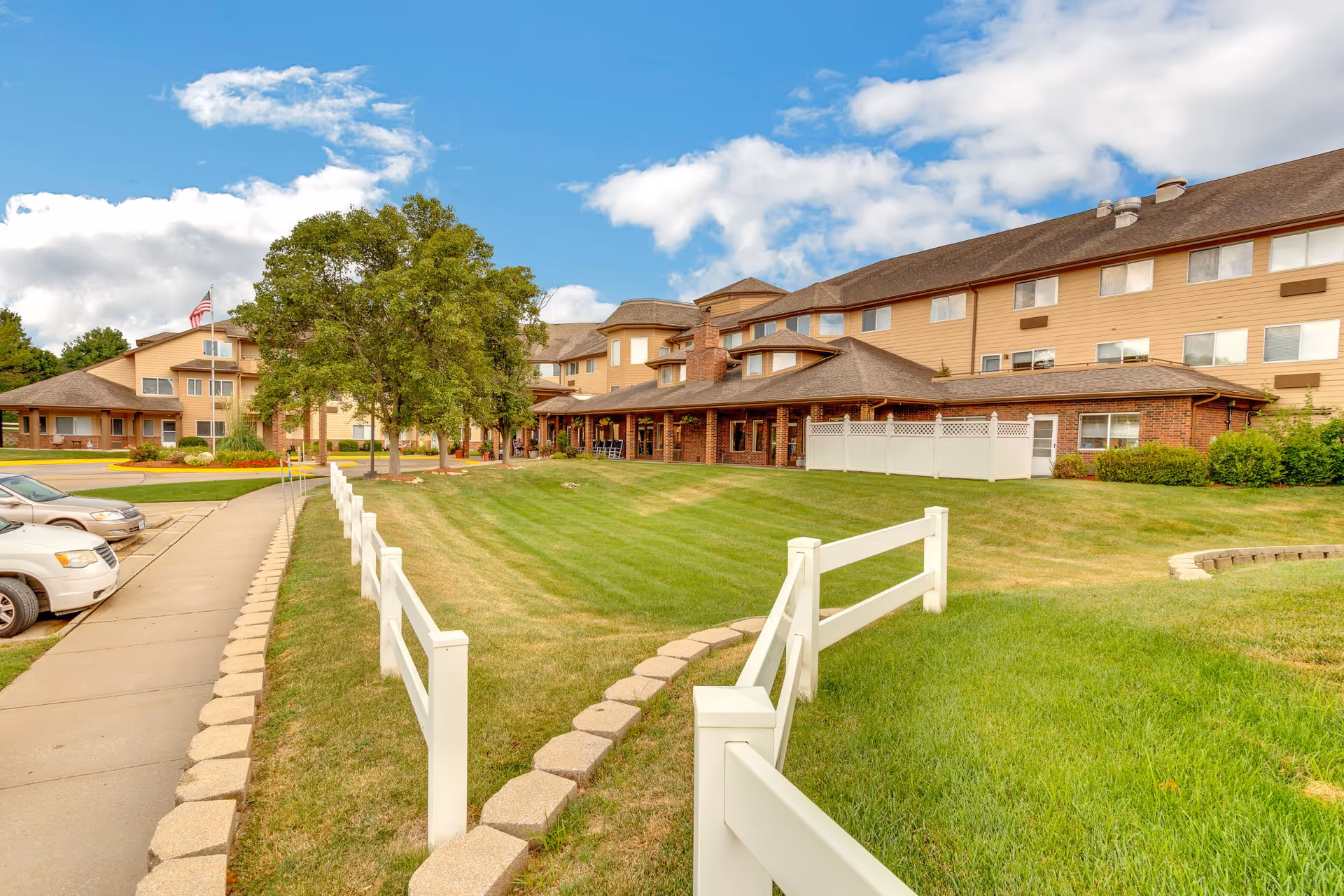 Front exterior of a multi-story senior living facility with a manicured lawn, white fence, and parked cars.