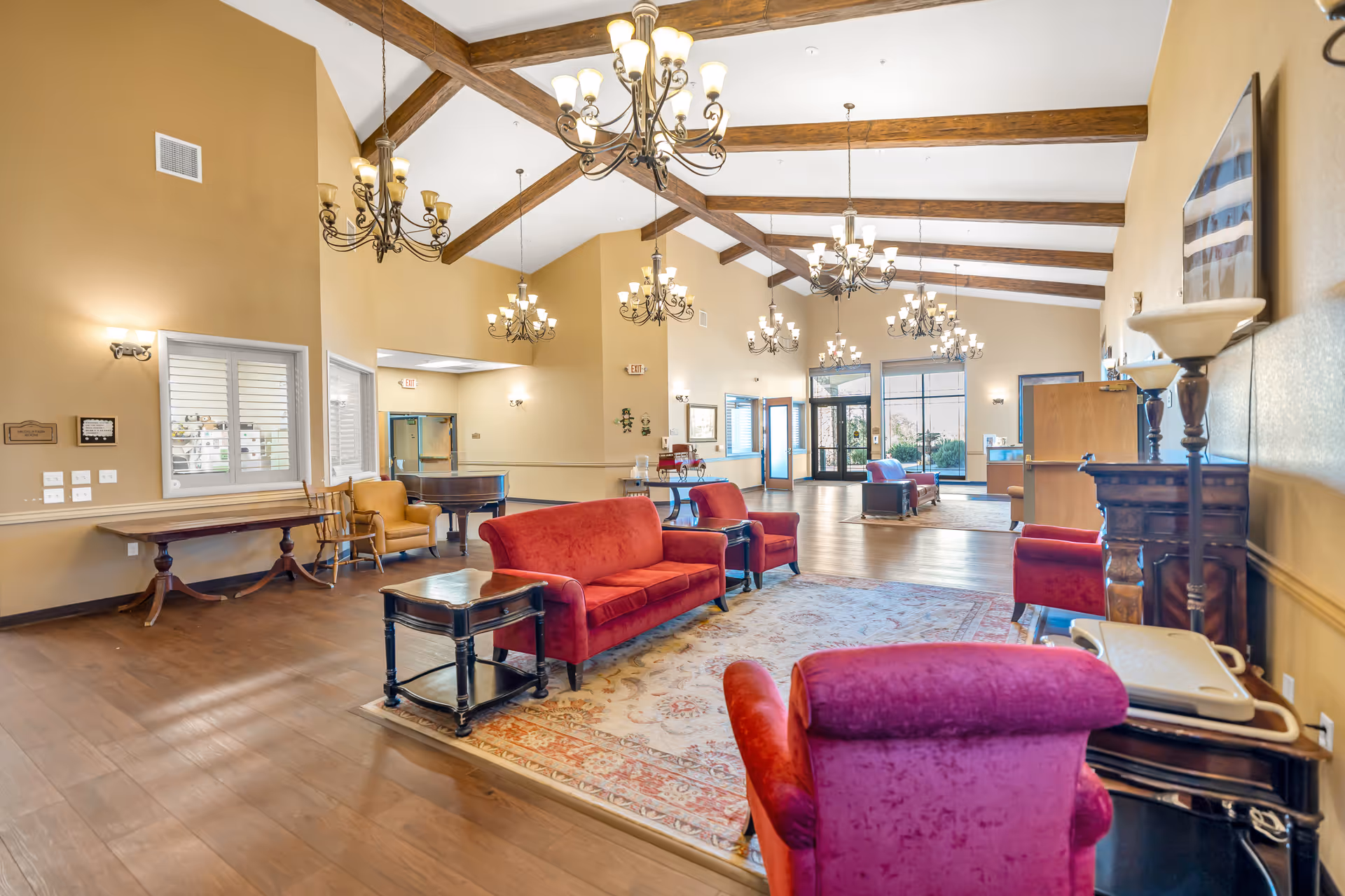 Spacious senior living facility common area with high vaulted ceilings featuring exposed wooden beams and multiple chandeliers. The room is furnished with red and purple upholstered sofas and armchairs arranged around a large patterned area rug. There is a wooden side table, a piano in the corner, and large windows and glass doors letting in natural light. The walls are painted beige with wooden trim, and the floor is hardwood.