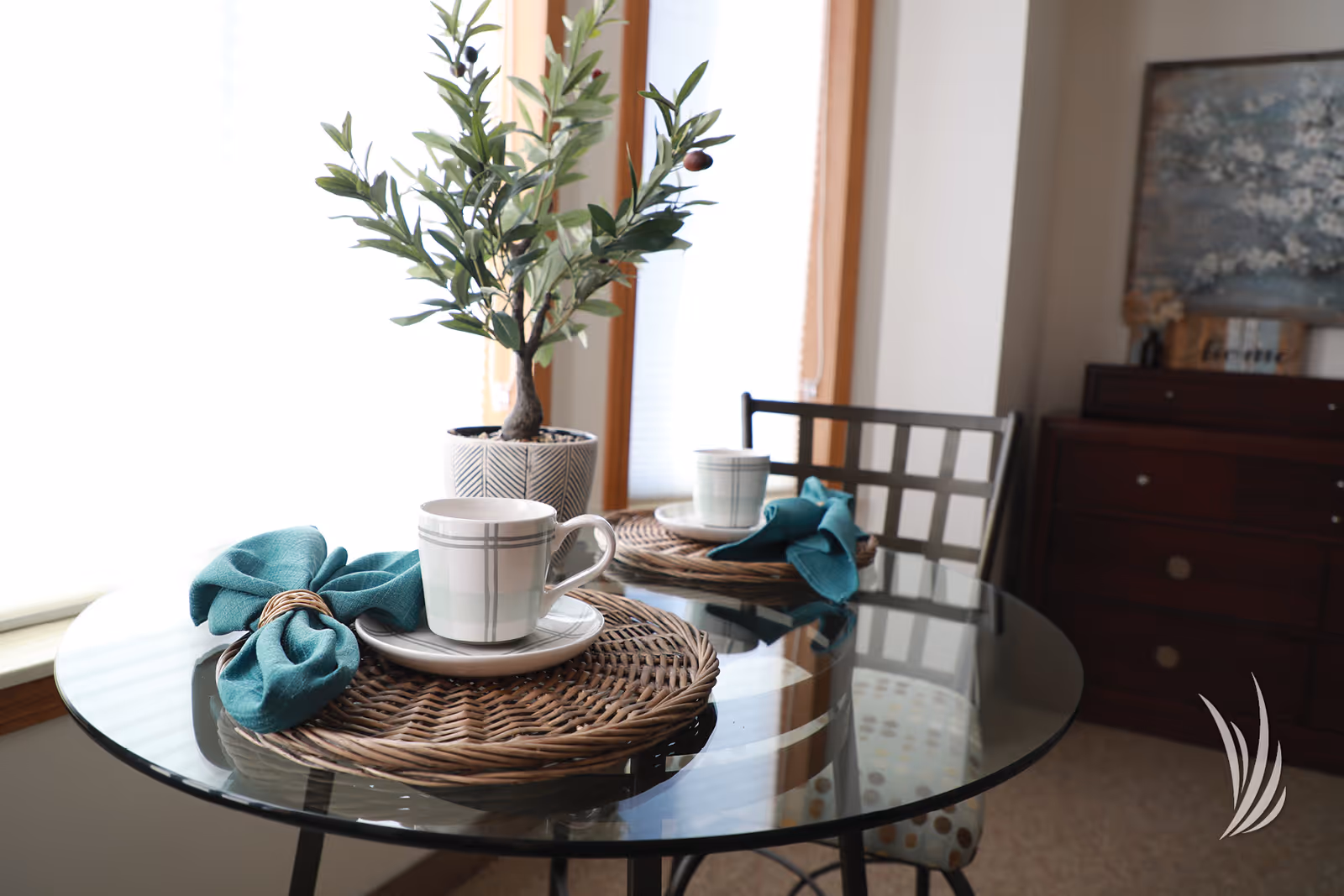 A glass-top dining table set for two with cups, teal napkins, woven placemats and a potted plant centerpiece in a bright dining area.