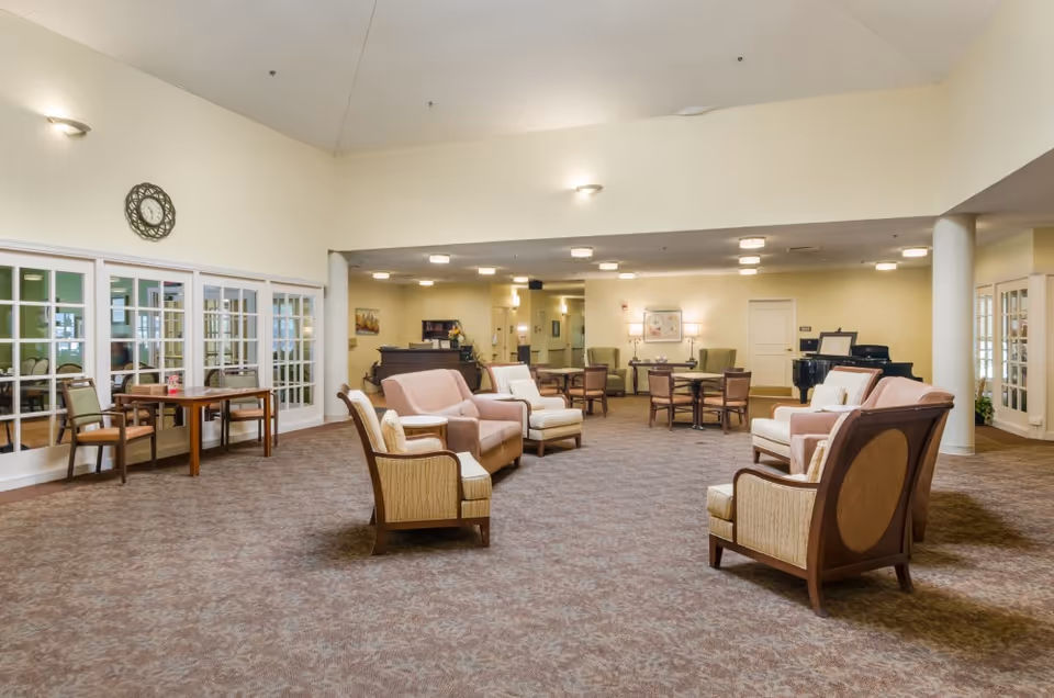 A spacious senior living facility common area with multiple seating arrangements including armchairs and sofas. The room has beige walls, carpeted floors, and soft lighting. There are tables with chairs along the walls, a clock on one wall, and a piano in the background. Large windows with glass panes separate this room from an adjacent room.