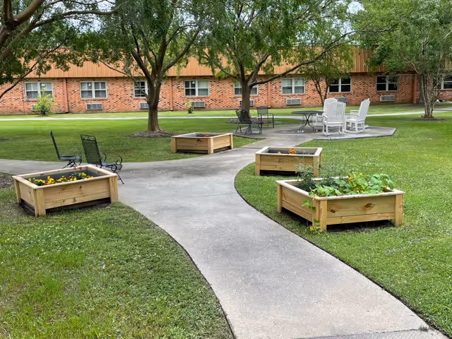 Outdoor garden area with a concrete pathway winding through raised wooden planter boxes containing flowers and plants. Several trees provide shade, and there are black metal benches and white chairs around a small paved seating area. A brick building with multiple windows is visible in the background.