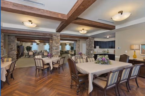 A spacious dining area in a senior living facility with multiple tables covered in beige tablecloths, surrounded by chairs with striped cushions. The room features wooden beams on the ceiling, stone pillars, and a wooden floor with a herringbone pattern. In the background, there is a kitchenette area with dark cabinets and a countertop, and the space is well-lit with ceiling lights and a table lamp.