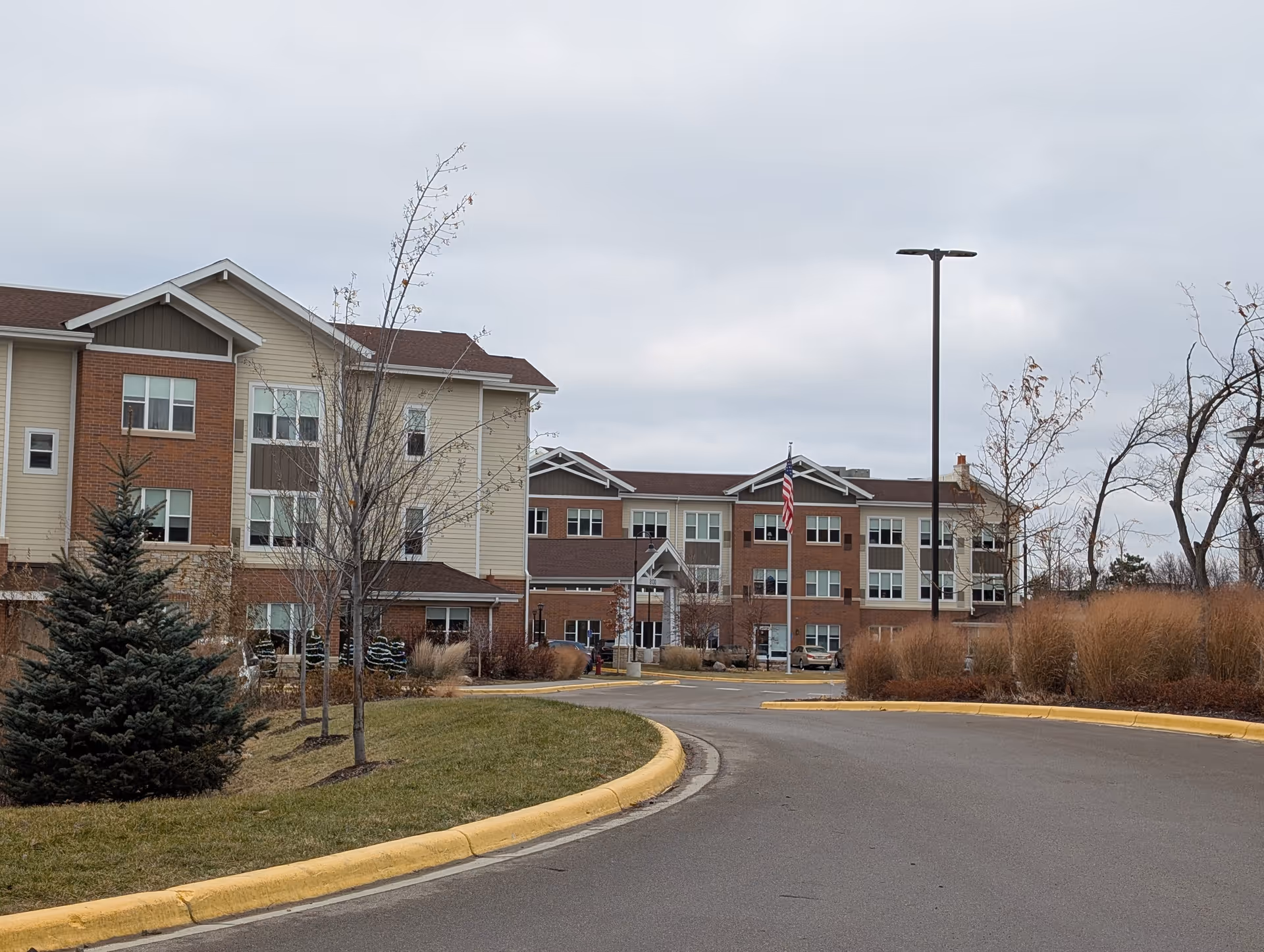 Exterior view of a multi-story senior living facility building with beige and brick facade, surrounded by landscaped grass, trees, and shrubs under a cloudy sky. A curved driveway with a yellow curb leads to the entrance, where an American flag is displayed on a flagpole.