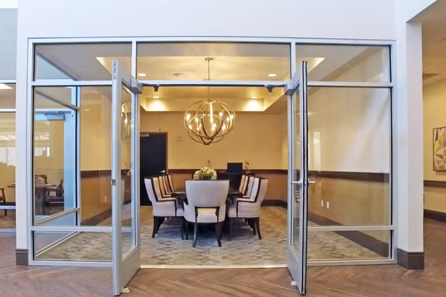 A conference or meeting room with glass double doors open, showing a rectangular table surrounded by eight upholstered chairs. A modern spherical chandelier hangs above the table, and a floral centerpiece is placed in the middle. The room has beige walls and carpeted flooring.