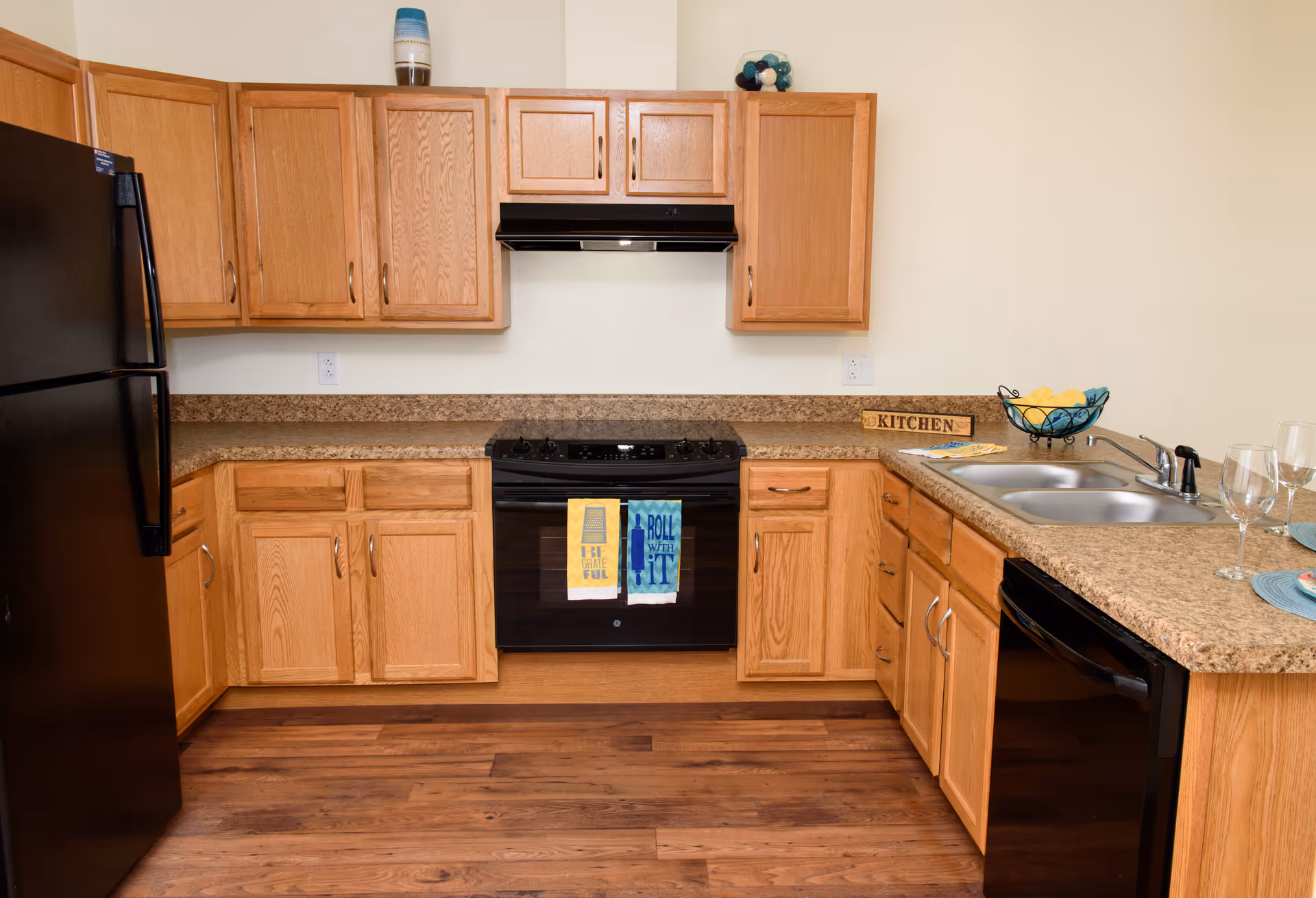 A kitchen with wooden cabinets, a black refrigerator, a black stove with two colorful towels hanging on the handle, a double sink, a dishwasher, and a countertop with a decorative bowl of lemons and two wine glasses.