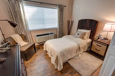 A cozy bedroom with a single bed covered in beige bedding, a wooden nightstand with a lamp and books, a cushioned chair with a throw blanket, a window with closed blinds and curtains, and wooden flooring with a small rug beside the bed.