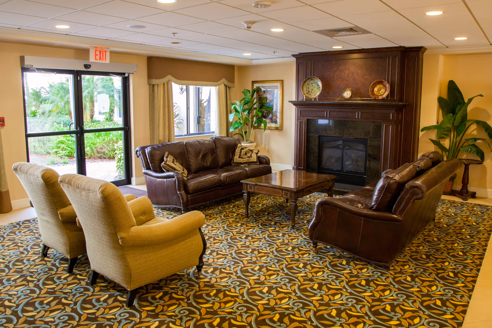 A cozy living room area with two brown leather sofas and two beige armchairs arranged around a wooden coffee table on a patterned carpet. There is a dark wooden fireplace with decorative plates and a clock on the mantel. Large windows and a glass door provide a view of greenery outside. Potted plants and framed artwork decorate the room.