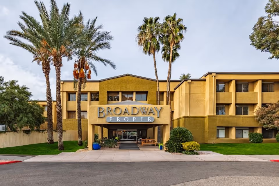 Front exterior view of Broadway Proper Assisted & Senior Living facility with a three-story building, palm trees, green lawn, and a covered entrance with the facility name displayed.
