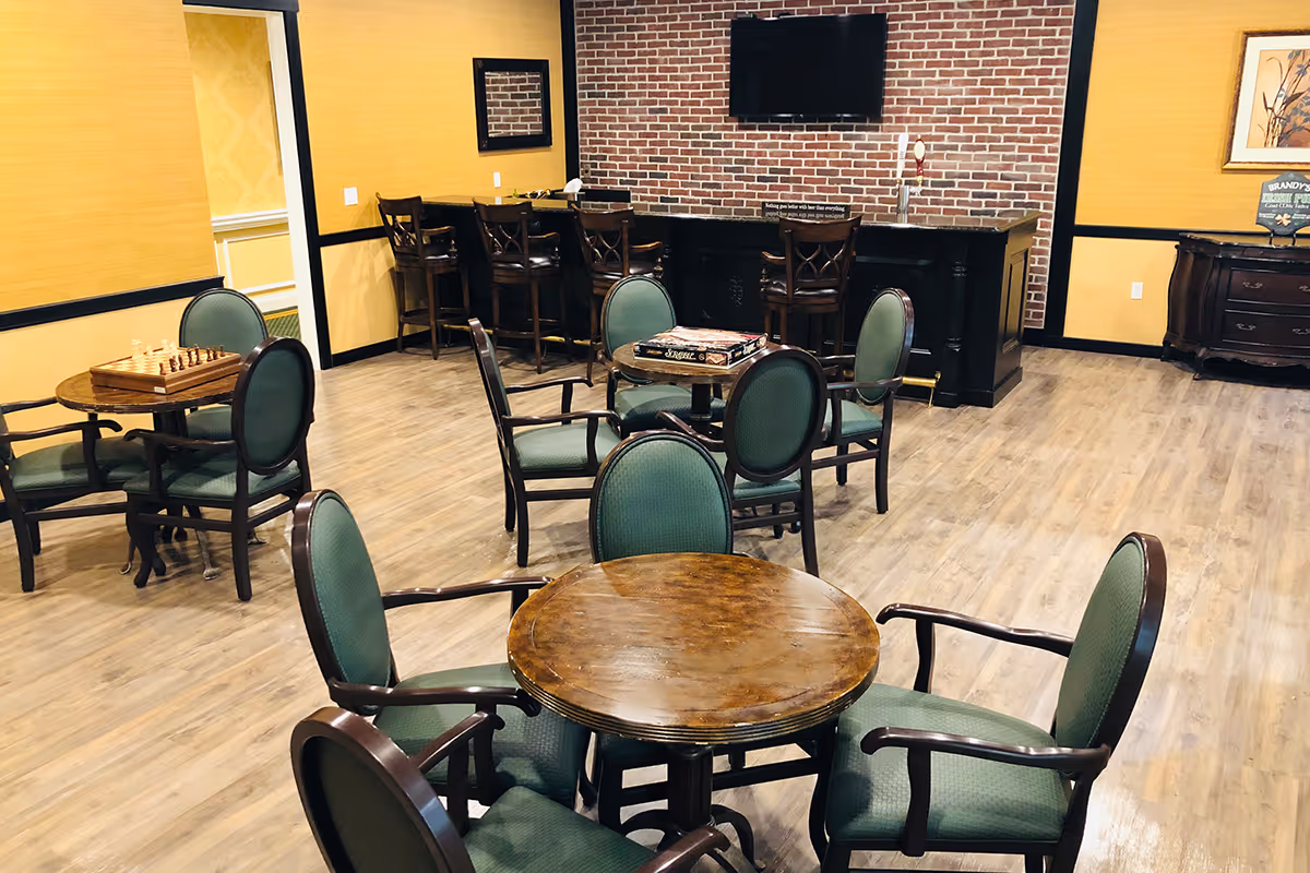 A common area with several round wooden tables surrounded by green cushioned chairs. One table has a chessboard, and another has a board game. In the background, there is a bar counter with high chairs and a flat-screen TV mounted on a brick wall. The room has wood flooring and yellow walls with black trim.