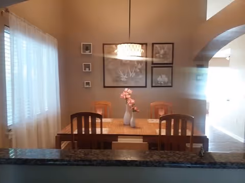 Dining room with a wooden table and four chairs, a pendant light, wall art, and a vase of flowers, viewed from behind a granite countertop.