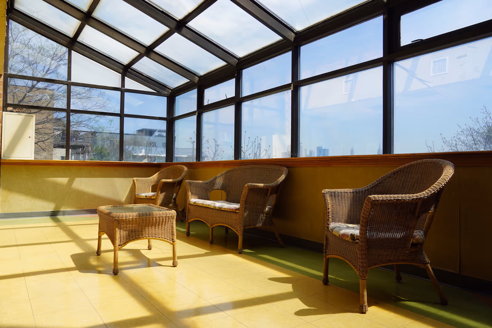 Sunlit enclosed sitting area with wicker chairs and a small wicker table under large glass windows and skylights.