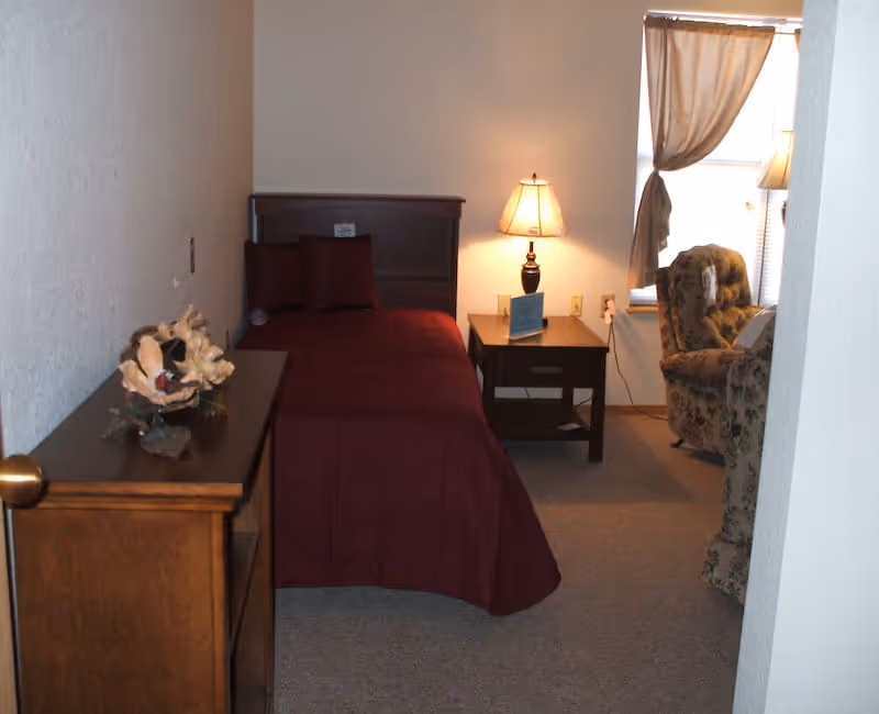 Small bedroom with a single bed dressed in a burgundy comforter, a bedside table with a lamp, a dresser, and an upholstered chair by a curtained window.