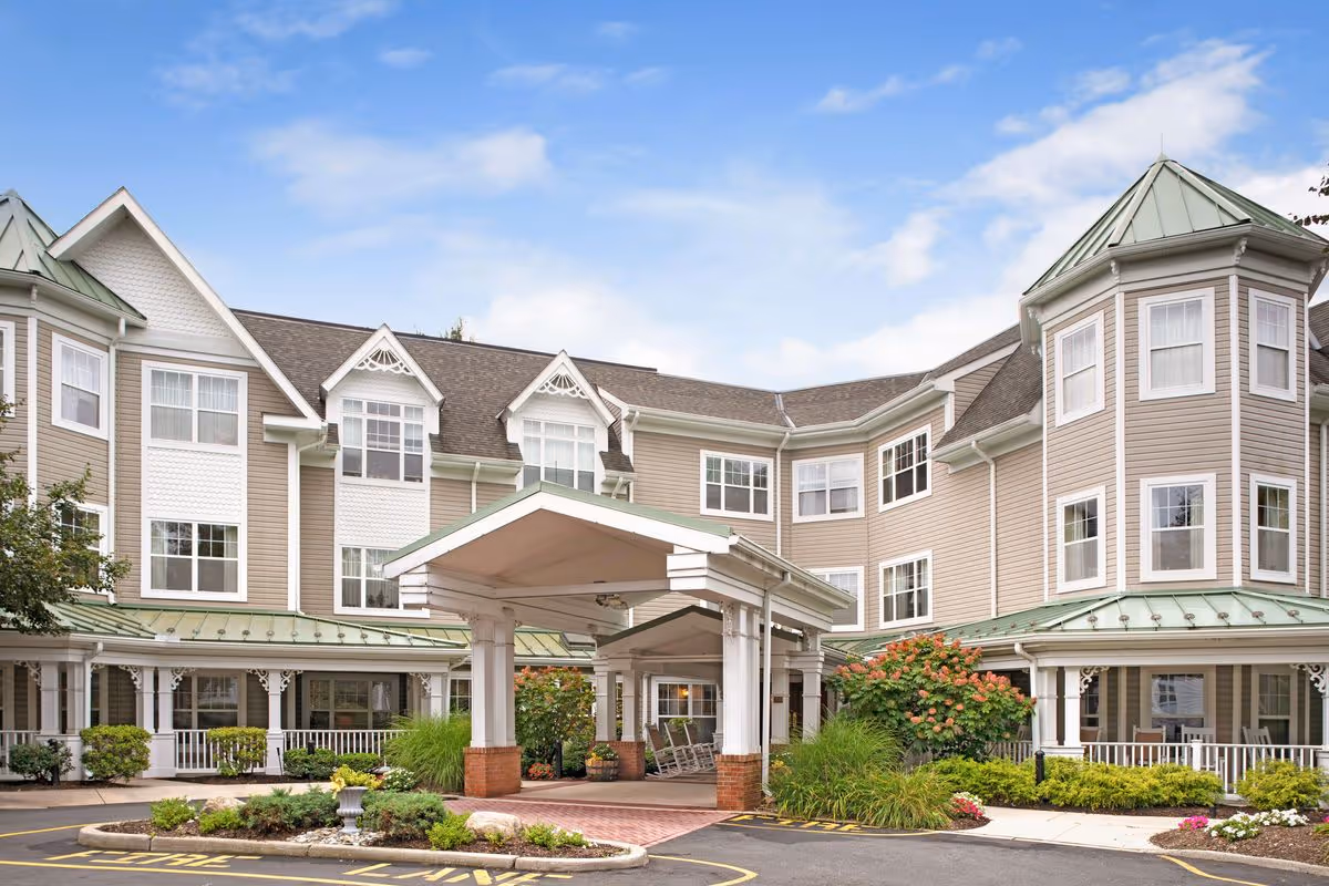 Exterior view of a senior living facility building with beige siding, white trim, and green metal roofs. The building has multiple windows and a covered entrance with columns. There are landscaped bushes and flowers around the entrance and a driveway with a fire lane marking.