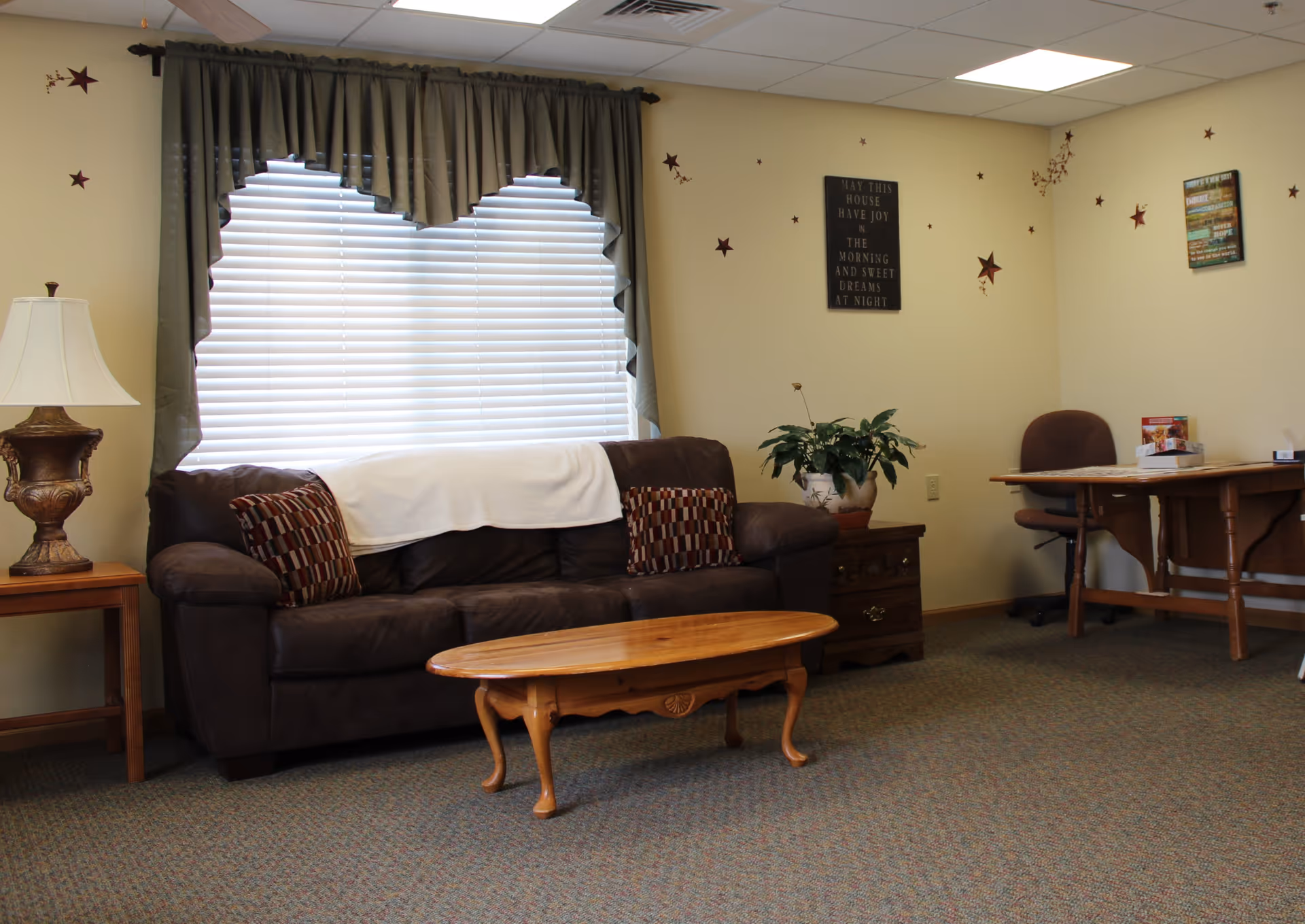 A cozy living room with a dark brown sofa adorned with two patterned cushions and a white throw draped over the back. In front of the sofa is a wooden coffee table. To the left of the sofa is a side table with a decorative lamp. The window behind the sofa has white blinds and green curtains. On the right side of the room, there is a wooden desk with a brown office chair and some items on the desk. The walls are decorated with star-shaped decals and framed inspirational quotes. A potted plant sits on a small wooden cabinet next to the sofa.