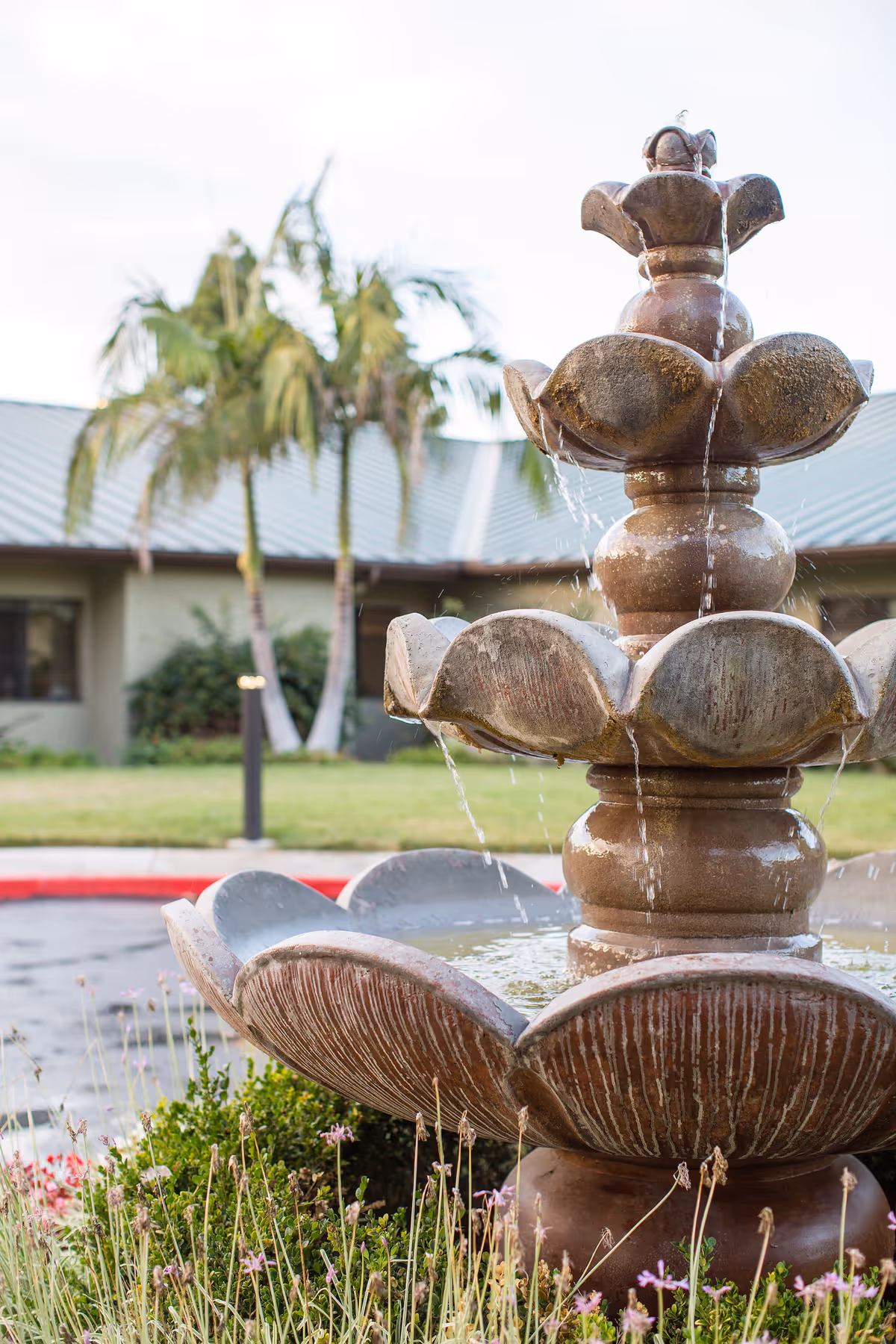 A tiered stone fountain with water flowing in front of a one-story building and palm trees.