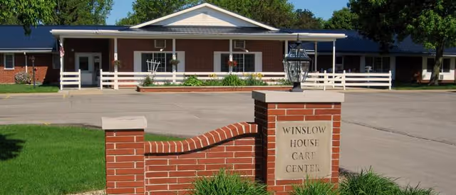 Front exterior view of Winslow House Care Center, a single-story brick building with a white roof trim and a covered entrance. In front of the building is a brick sign with the facility's name, Winslow House Care Center, surrounded by green grass and a paved driveway.