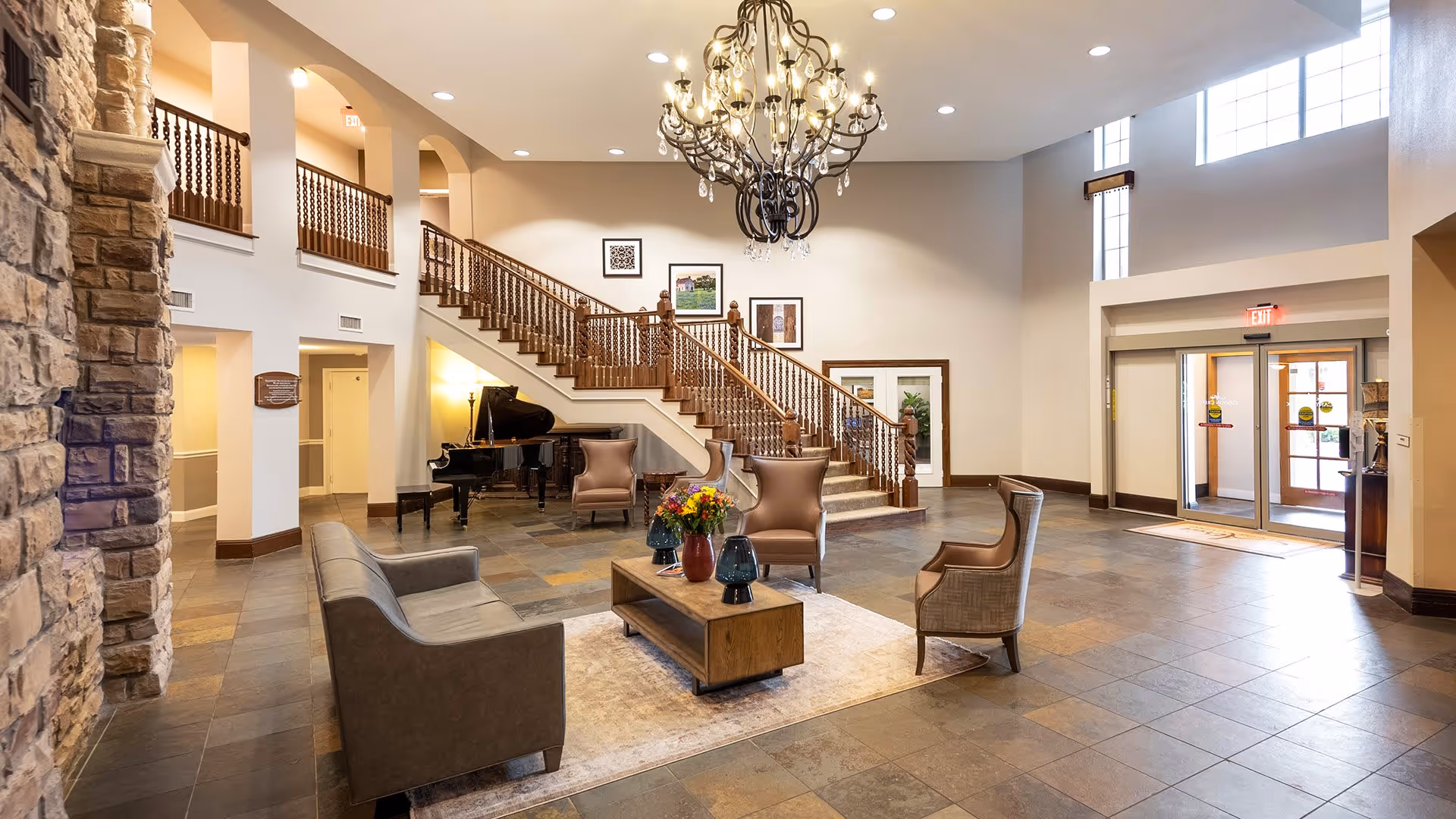 Spacious and well-lit senior living facility lobby with a large chandelier, stone accent wall, wooden staircase with ornate railing, a grand piano, and comfortable seating including a sofa and armchairs arranged around a coffee table with flower vases.