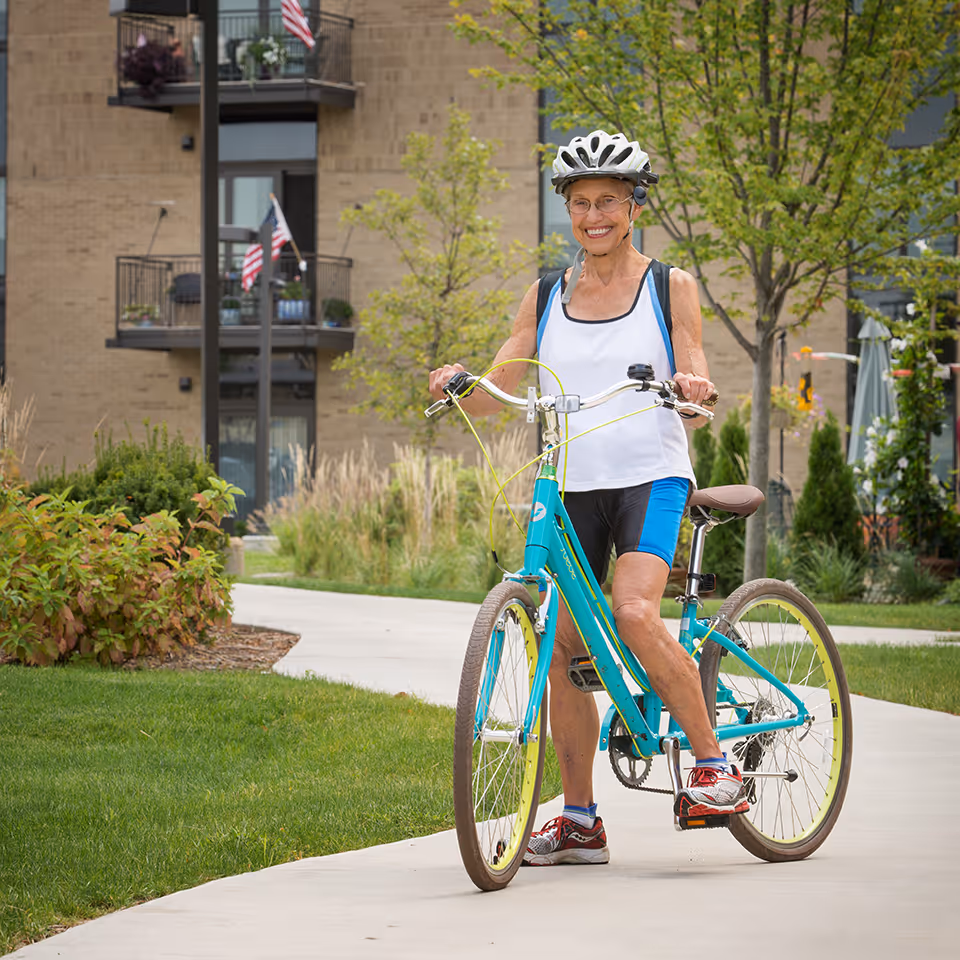 An elderly woman wearing a white helmet, white tank top, and blue cycling shorts is standing with a teal bicycle on a paved path in a landscaped outdoor area with grass, bushes, and trees. Behind her is a multi-story building with balconies and American flags.