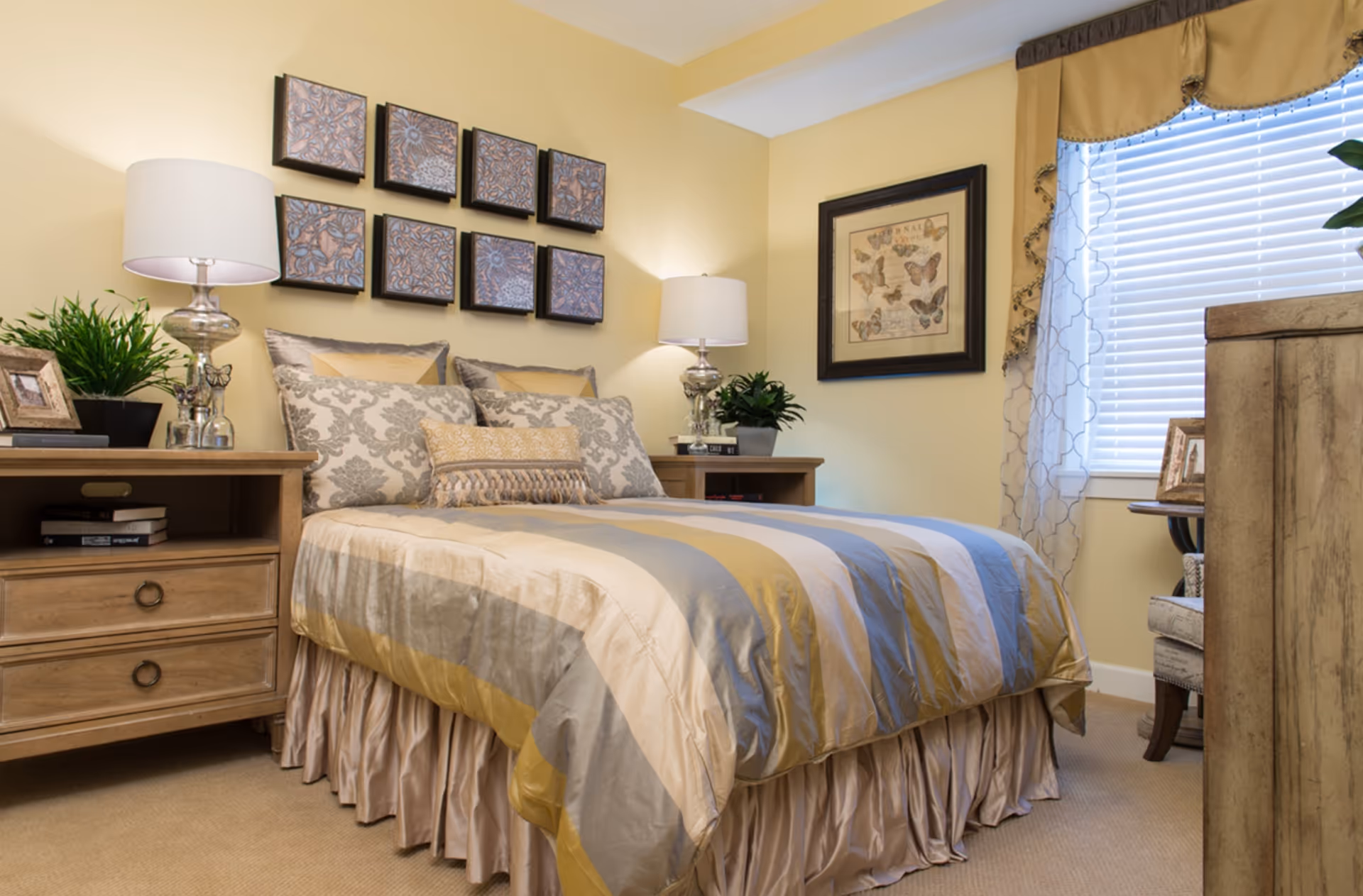 A cozy bedroom with a bed dressed in a striped comforter in shades of gold, gray, and beige. The bed has multiple decorative pillows and a ruffled bed skirt. On either side of the bed are wooden nightstands with table lamps, potted plants, and picture frames. Above the bed, there are nine square decorative wall art pieces arranged in a grid. A window with blinds and sheer curtains is on the right side of the room, next to a framed butterfly artwork on the wall. The room has soft yellow walls and beige carpeting.