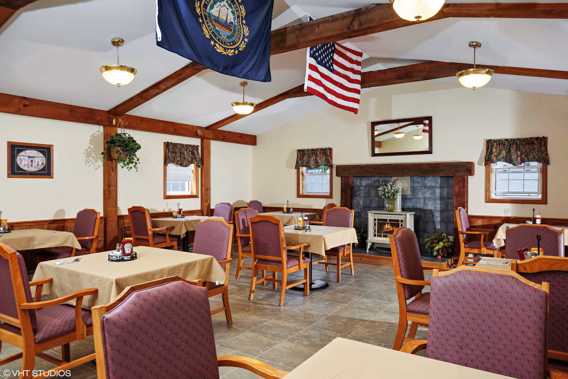 Dining room with multiple tables covered in beige tablecloths and surrounded by wooden chairs with purple cushions. The room features a fireplace with a small stove, a mirror above it, and two windows with floral valances. The ceiling has exposed wooden beams with hanging light fixtures and two flags, one American and one state flag.