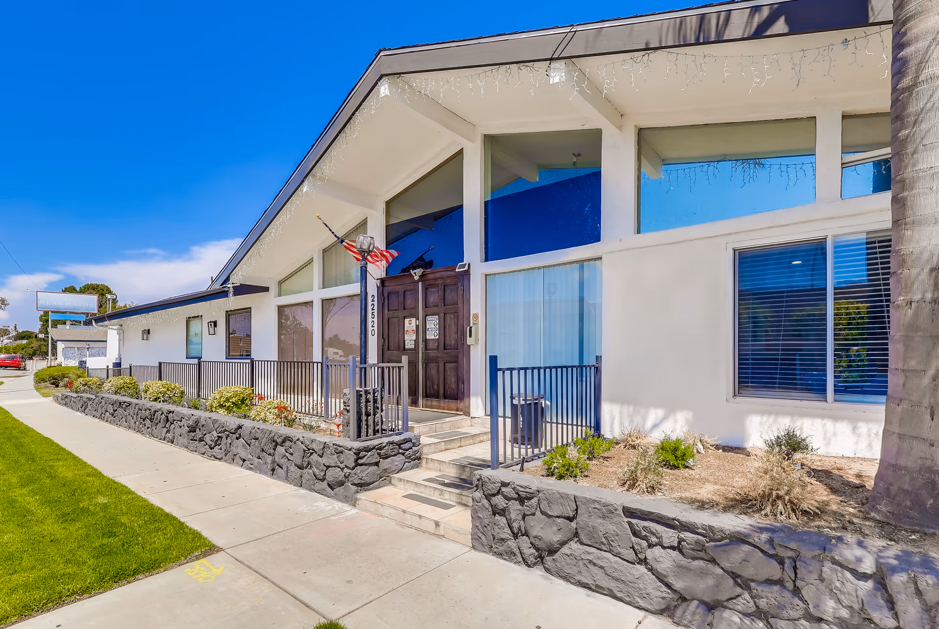 Exterior view of a single-story building with large windows and a dark wooden double door entrance. The building has a slanted roof with white icicle-style string lights hanging from the edge. There is a small stone wall with plants in front of the building and a sidewalk running alongside the building. An American flag is mounted near the entrance.
