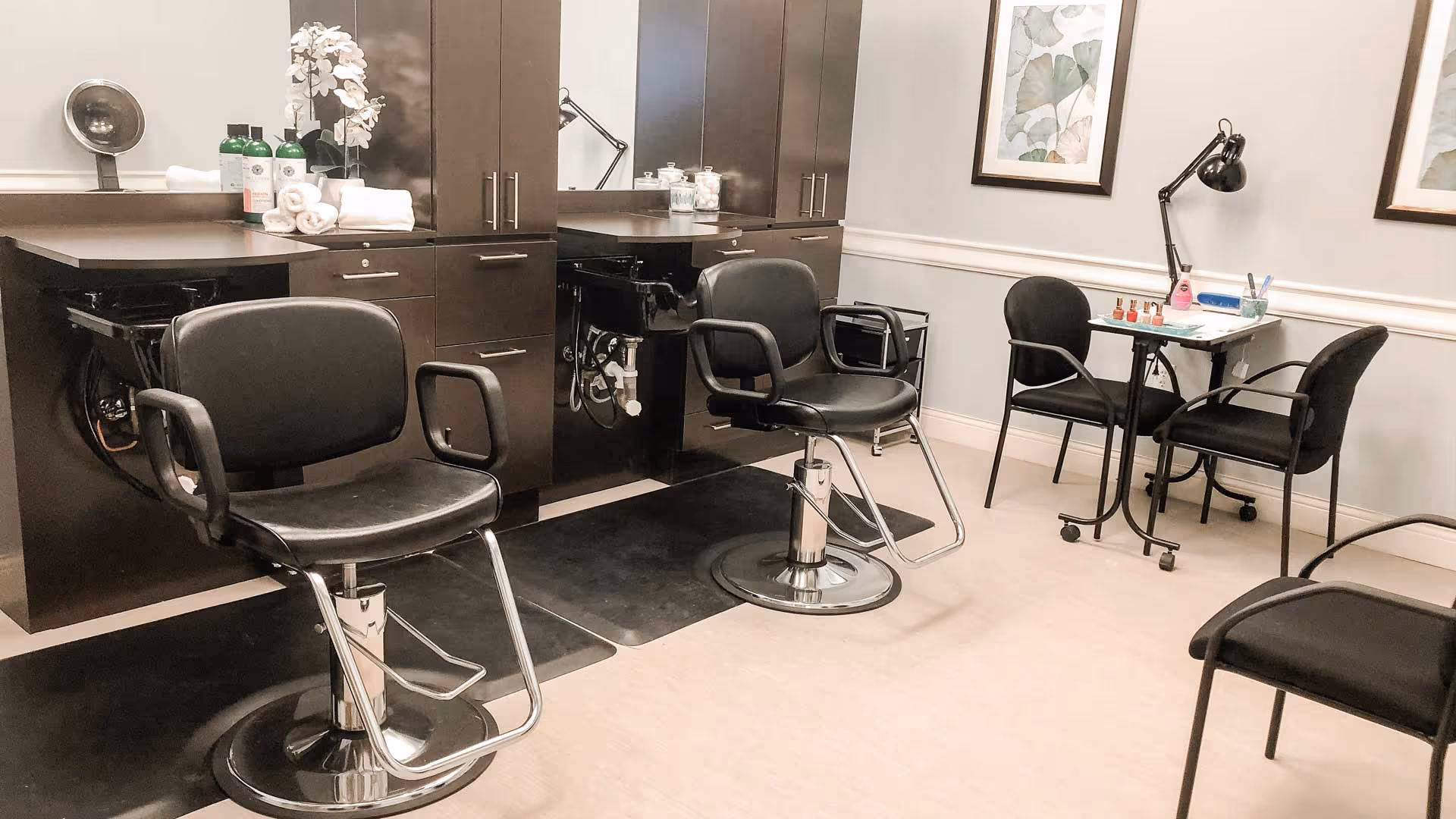 Small salon room with two black styling chairs in front of mirrored cabinets and a manicure table with chairs to the right.